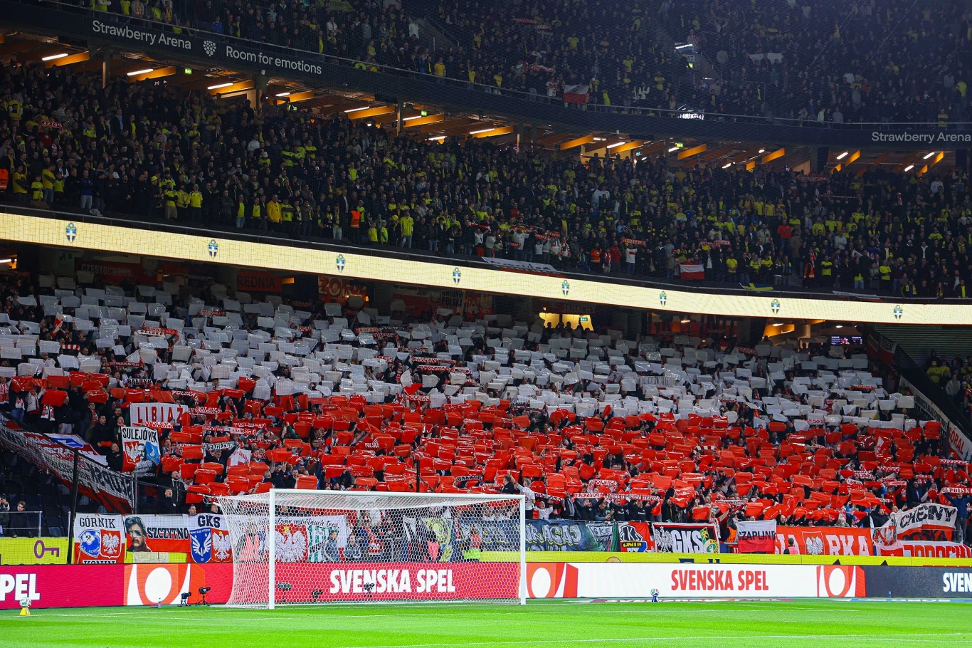 kibice ubrani na biało-czerwono tworzą ogromną flagę Polski na trybunach stadionu podczas meczu piłki nożnej, dominują szaliki, chorągiewki oraz kartoniki w barwach narodowych, a na murawie widoczna jest bramka i reklamy sponsorskie