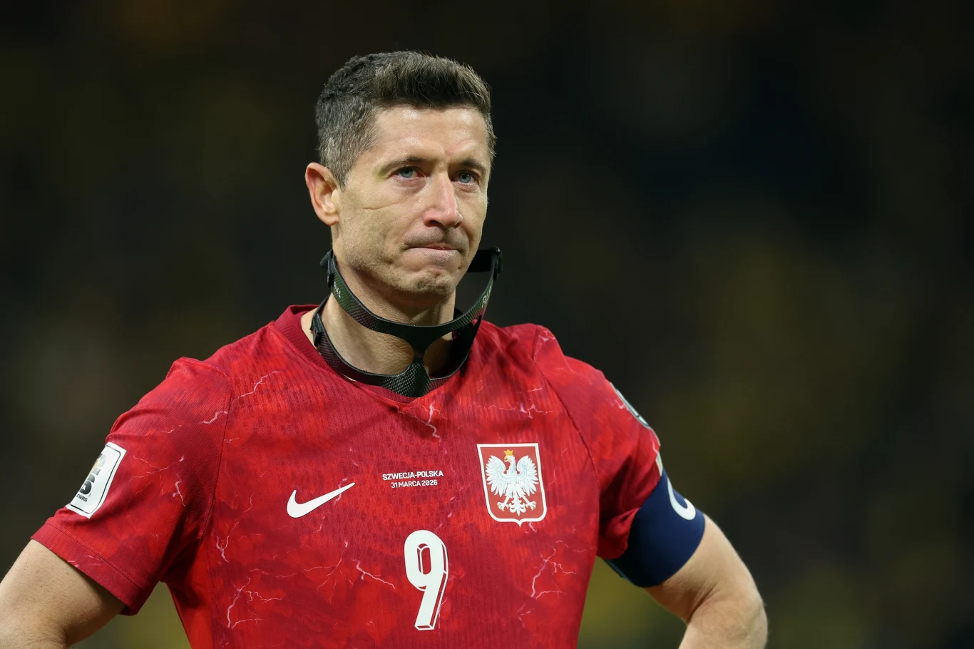 piłkarz w czerwonej koszulce reprezentacji Polski z numerem 9 i opaską kapitańską na ramieniu stoi na boisku z poważnym wyrazem twarzy