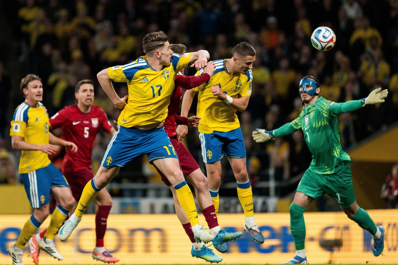 dynamiczna akcja piłkarska z udziałem zawodników w żółto-niebieskich strojach, którzy walczą o piłkę głową w polu karnym, podczas gdy bramkarz w zielonym stroju próbuje obronić strzał, wokół nich widać skupionych graczy obu drużyn