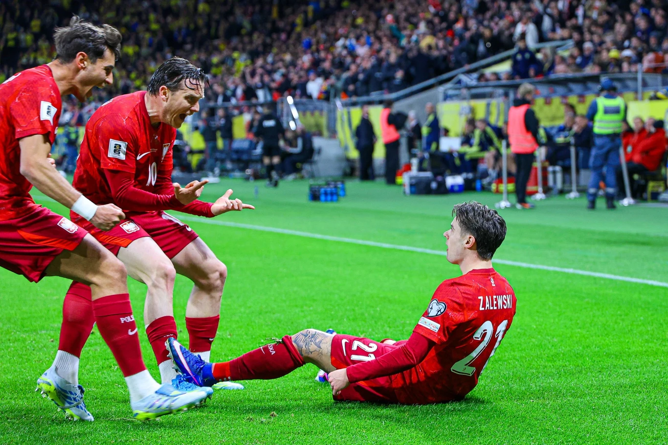 Piłkarz w czerwonej koszulce siedzi na murawie i świętuje zdobycie gola, podczas gdy dwaj koledzy z drużyny podbiegają do niego z szerokimi uśmiechami, z tyłu widać trybuny pełne kibiców oraz obsługę techniczną przy linii bocznej boiska.