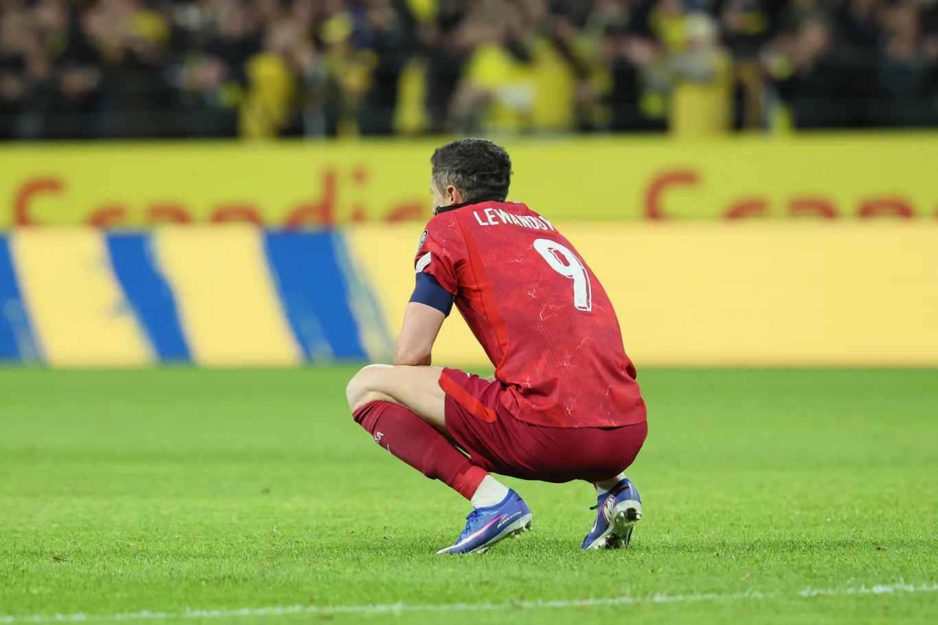 piłkarz w czerwonym stroju z numerem 9 i nazwiskiem na plecach kuca na murawie stadionu, w tle kibice oraz żółte trybuny