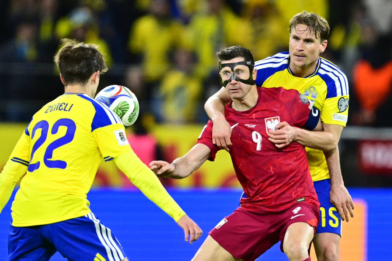 piłkarz w czerwonym stroju z numerem 9 i czarną maską na twarzy prowadzi piłkę, naciskany przez zawodnika w żółto-niebieskiej koszulce z numerem 15; z lewej strony zawodnik z numerem 22 obserwuje akcję