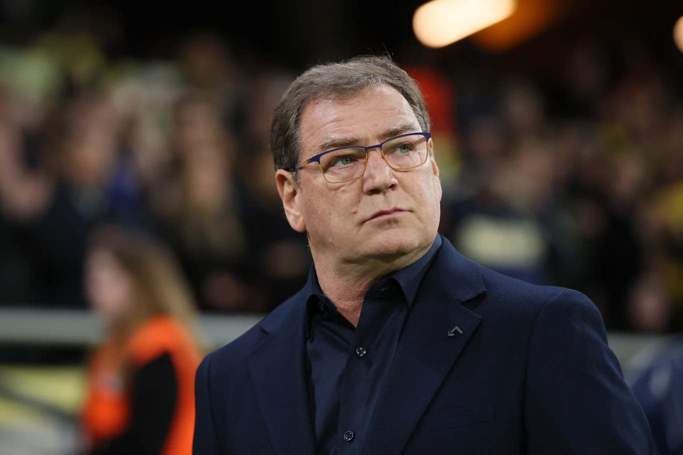 mężczyzna w okularach ubrany w ciemny garnitur i koszulę stoi na tle rozmytego tłumu, zwracając uwagę poważnym wyrazem twarzy