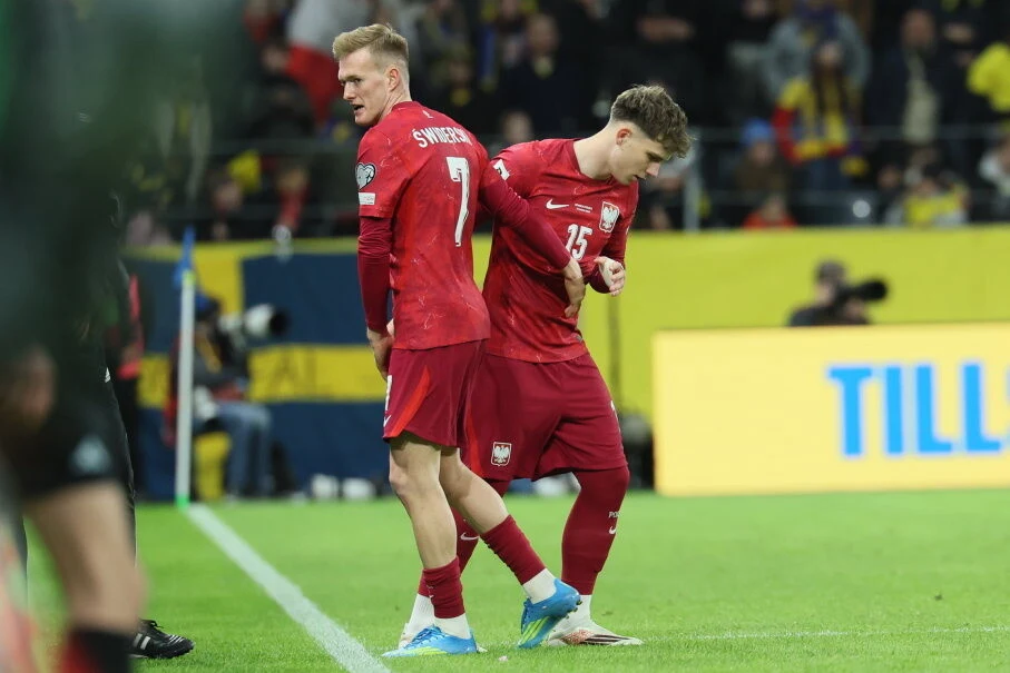 Dwóch piłkarzy w czerwonych strojach w trakcie zmiany na boisku, jeden z nich opuszcza murawę, drugi wchodzi do gry, w tle kibice oraz ławka rezerwowych.