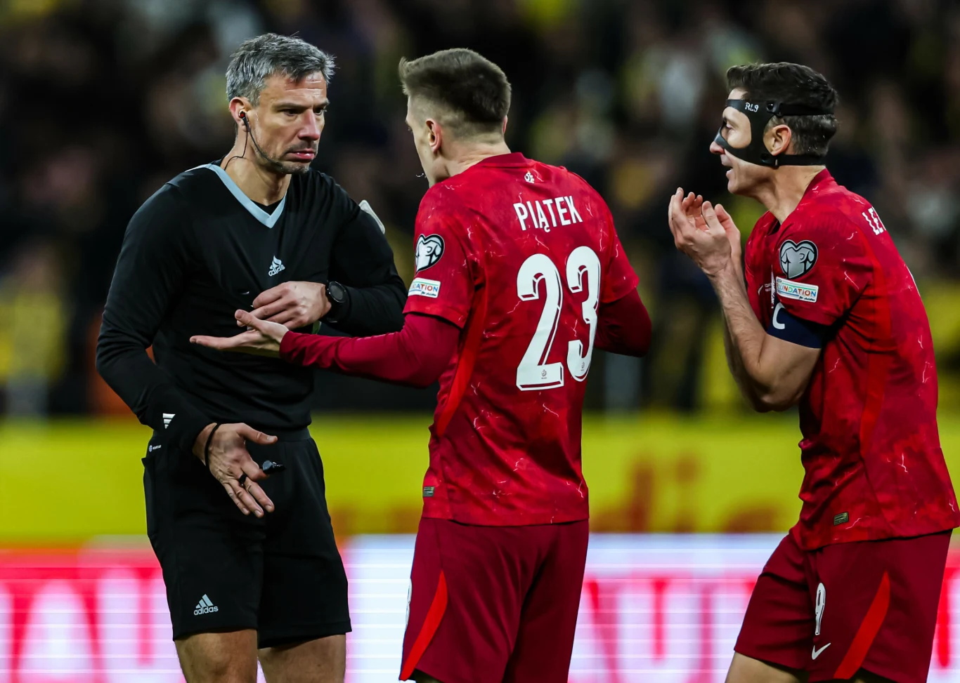 dwóch polskich piłkarzy w czerwonych strojach intensywnie gestykuluje w rozmowie z sędzią ubranym na czarno podczas meczu, w tle rozmyte postacie i żółte trybuny