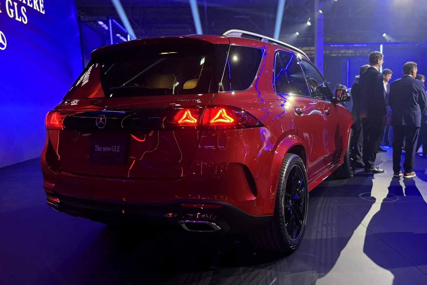 czerwony SUV marki Mercedes-Benz GLS ustawiony na ekspozycji, oświetlony reflektorami, z widocznymi czarnymi felgami i podświetlonymi tylnymi światłami; obok samochodu stoi kilku elegancko ubranych mężczyzn rozmawiających w grupie