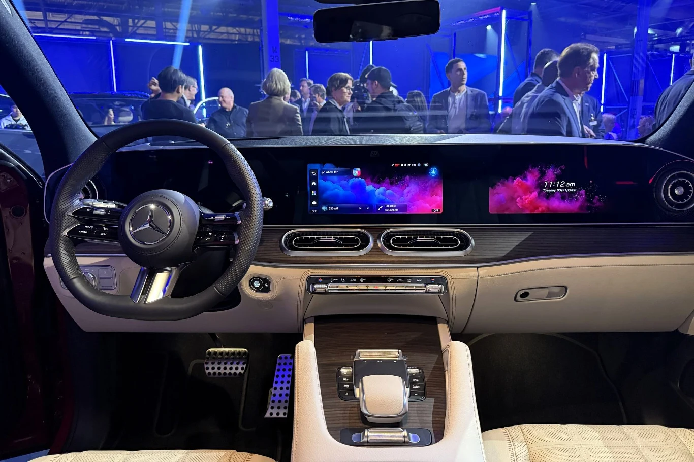 nowoczesne wnętrze luksusowego samochodu marki Mercedes z dużym, panoramicznym wyświetlaczem multimedialnym, elegancką deską rozdzielczą, wielofunkcyjną kierownicą i konsolą środkową wykończoną drewnem; w tle widoczni ludzie podczas prezentacji pojazdu