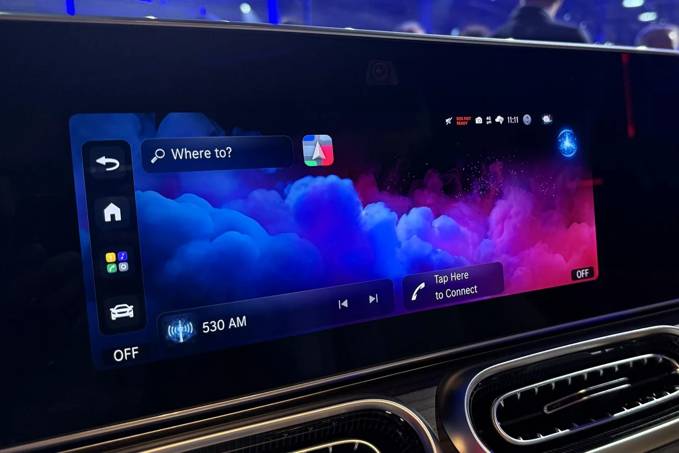 nowoczesny dotykowy ekran multimedialny w samochodzie wyświetlający kolorową grafikę z chmurami; widoczny panel nawigacji, ustawienia radia oraz sugestie połączeń z urządzeniem mobilnym