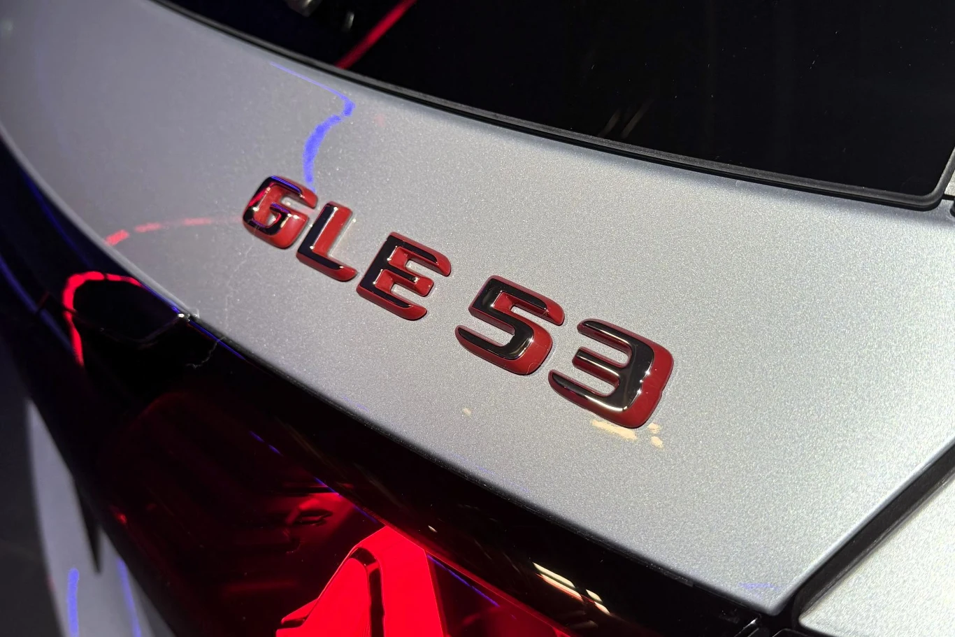 czerwony emblemat GLE 53 na srebrnej karoserii samochodu, podkreślony odbiciem świateł oraz połyskiem lakieru