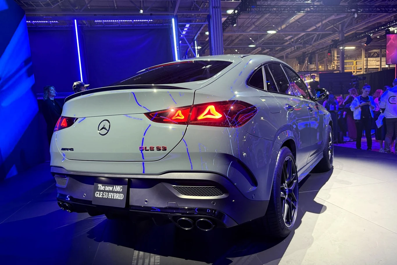 Srebrny Mercedes-AMG GLE53 HYBRID ustawiony na wystawie motoryzacyjnej, oświetlony fioletowym światłem, z wyraźnie widocznymi tylnymi światłami LED, podwójnymi końcówkami wydechu oraz detalami nadwozia typu coupe, otoczony grupą osób zwracających uwagę...