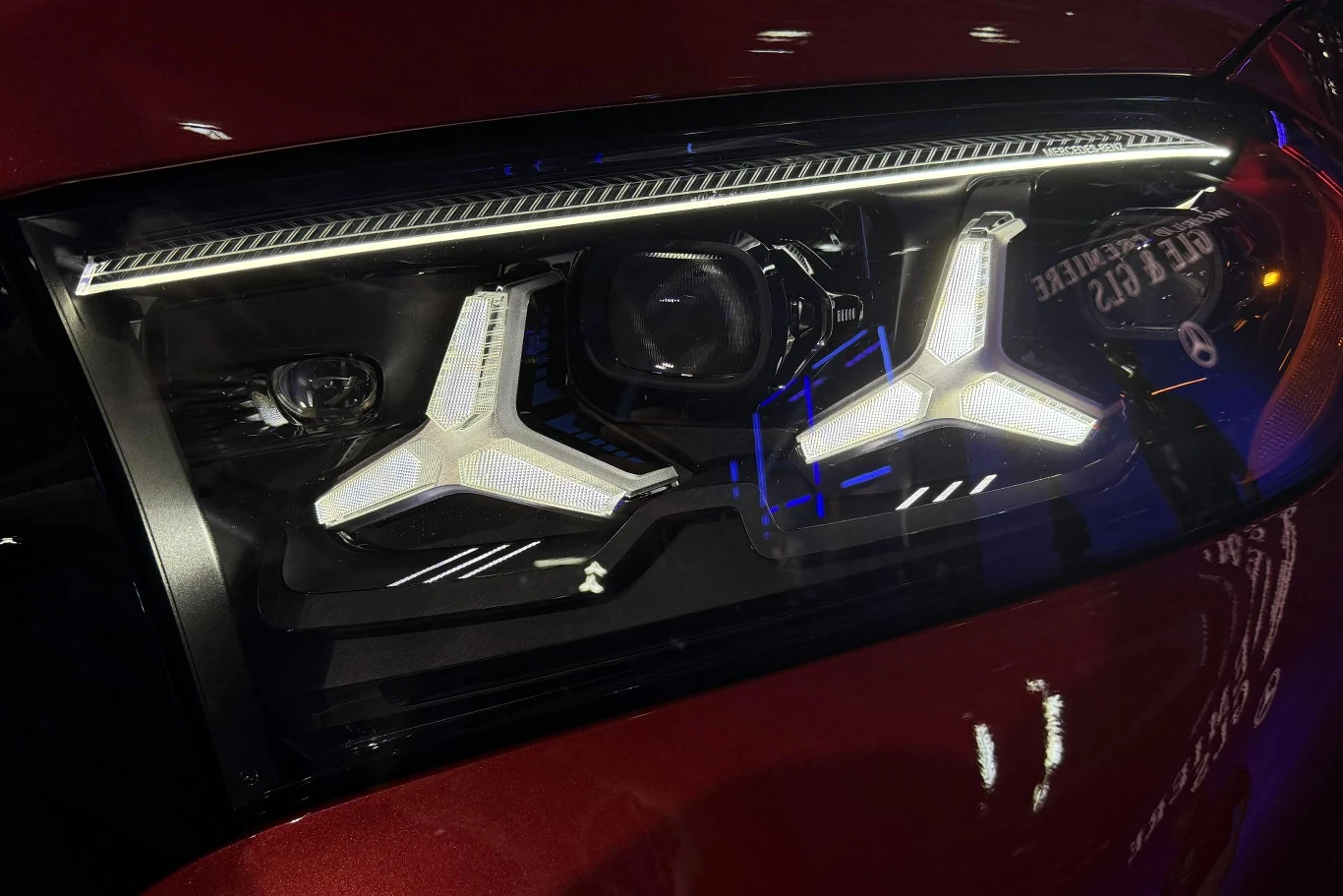 nowoczesny reflektor samochodowy z charakterystycznymi, trójramiennymi światłami LED umieszczonymi w czarnej oprawie, odbijający elementy otoczenia na błyszczącej, czerwonej karoserii