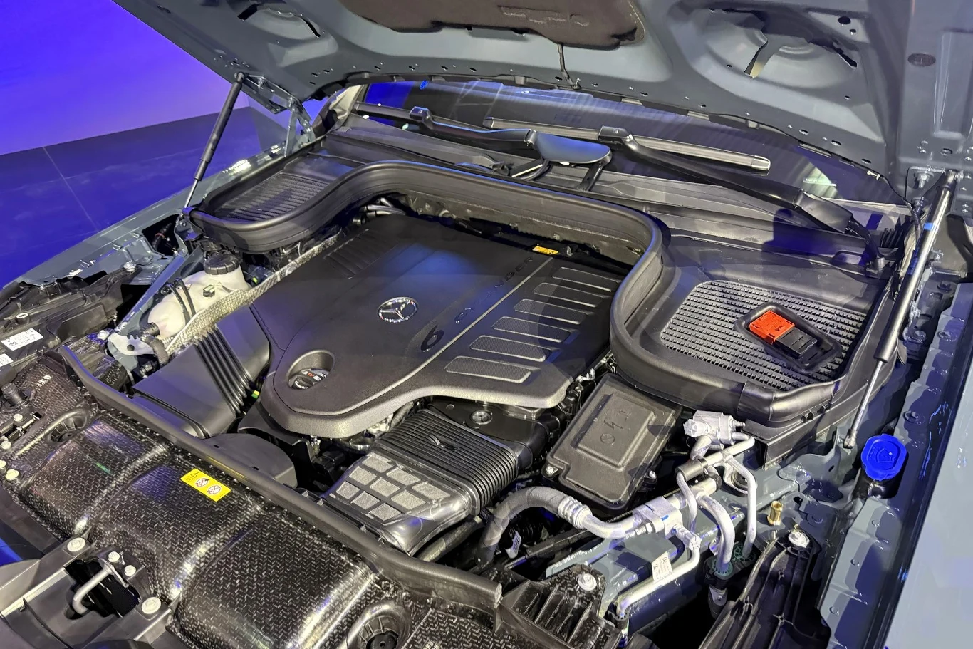 silnik samochodowy z logo Mercedesa, osłonięty nowoczesną plastikową pokrywą, z wyraźnie widocznymi elementami układu chłodzenia, przewodami technicznymi oraz solidnym mechanizmem mocowania maski