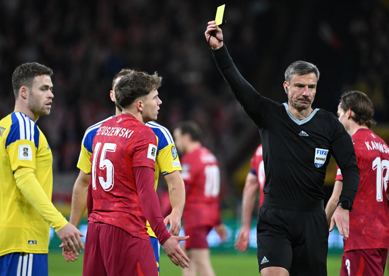 sędzia pokazuje żółtą kartkę piłkarzowi w czerwonym stroju, wokół znajdują się inni zawodnicy obu drużyn ubrane w czerwone i żółte stroje