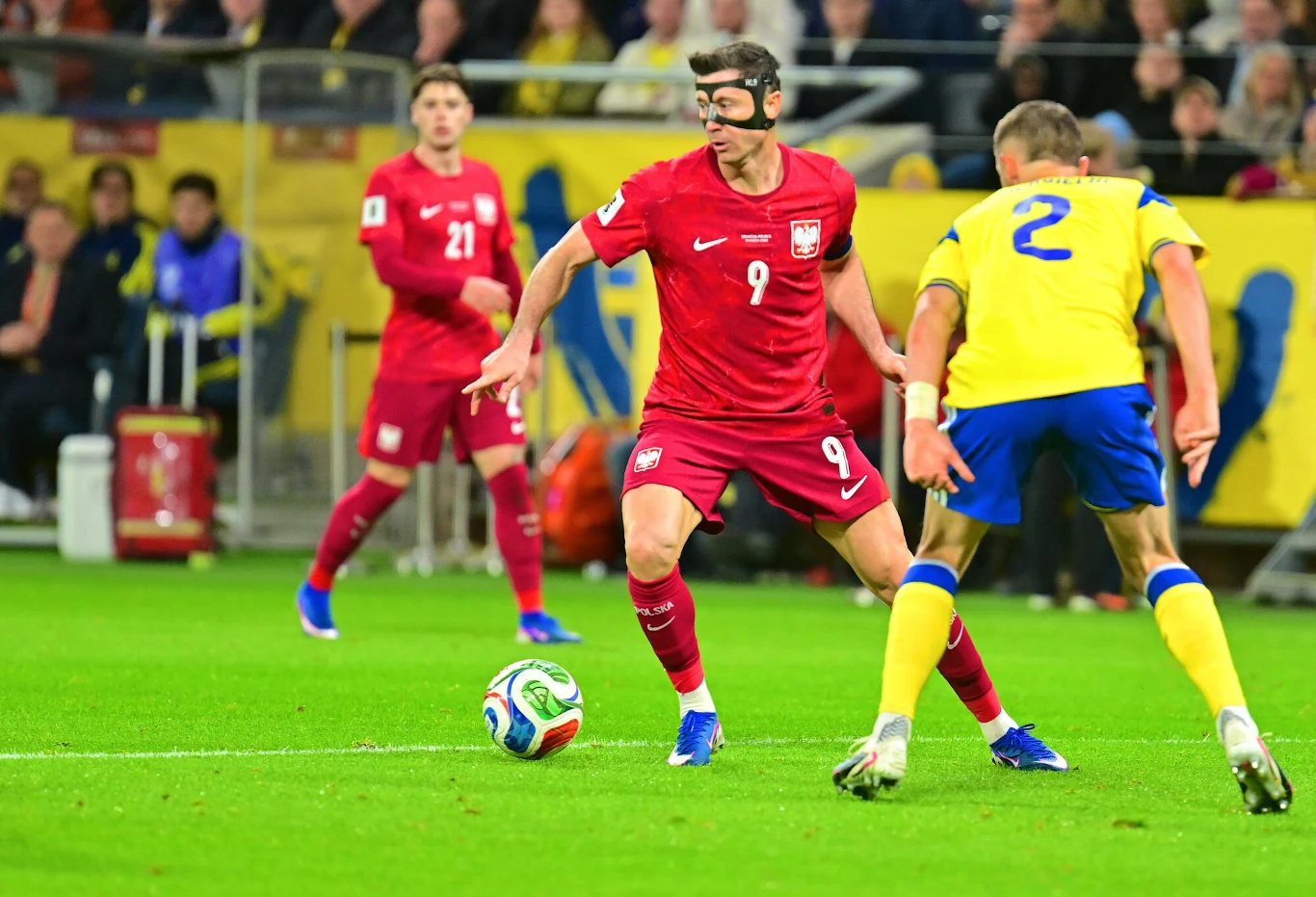 polski piłkarz w czerwonym stroju z opaską na głowie prowadzi piłkę podczas meczu przeciwko zawodnikowi w żółto-niebieskim stroju na zielonym boisku, w tle widoczni inni piłkarze i kibice na trybunach