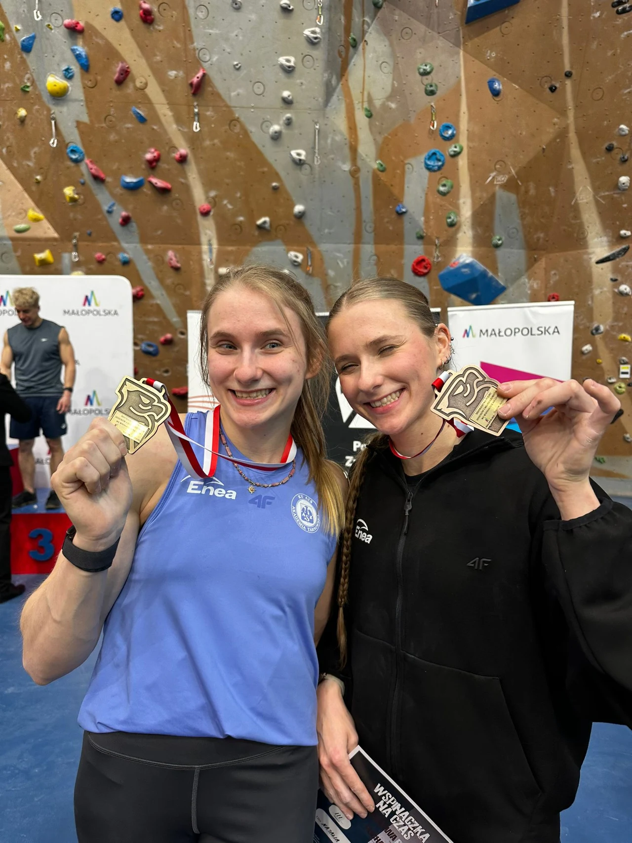 dwie uśmiechnięte kobiety stojące obok siebie na tle ściany wspinaczkowej trzymają w dłoniach złote medale, na szyjach mają zawieszone czerwono-białe wstążki, w tle widoczne podium oraz logo Małopolski