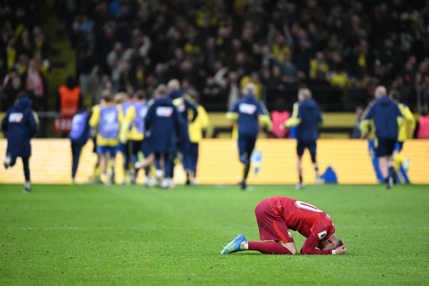 piłkarz w czerwonym stroju klęczący na murawie ze spuszczoną głową, podczas gdy w tle drużyna w żółto-niebieskich strojach cieszy się ze zwycięstwa