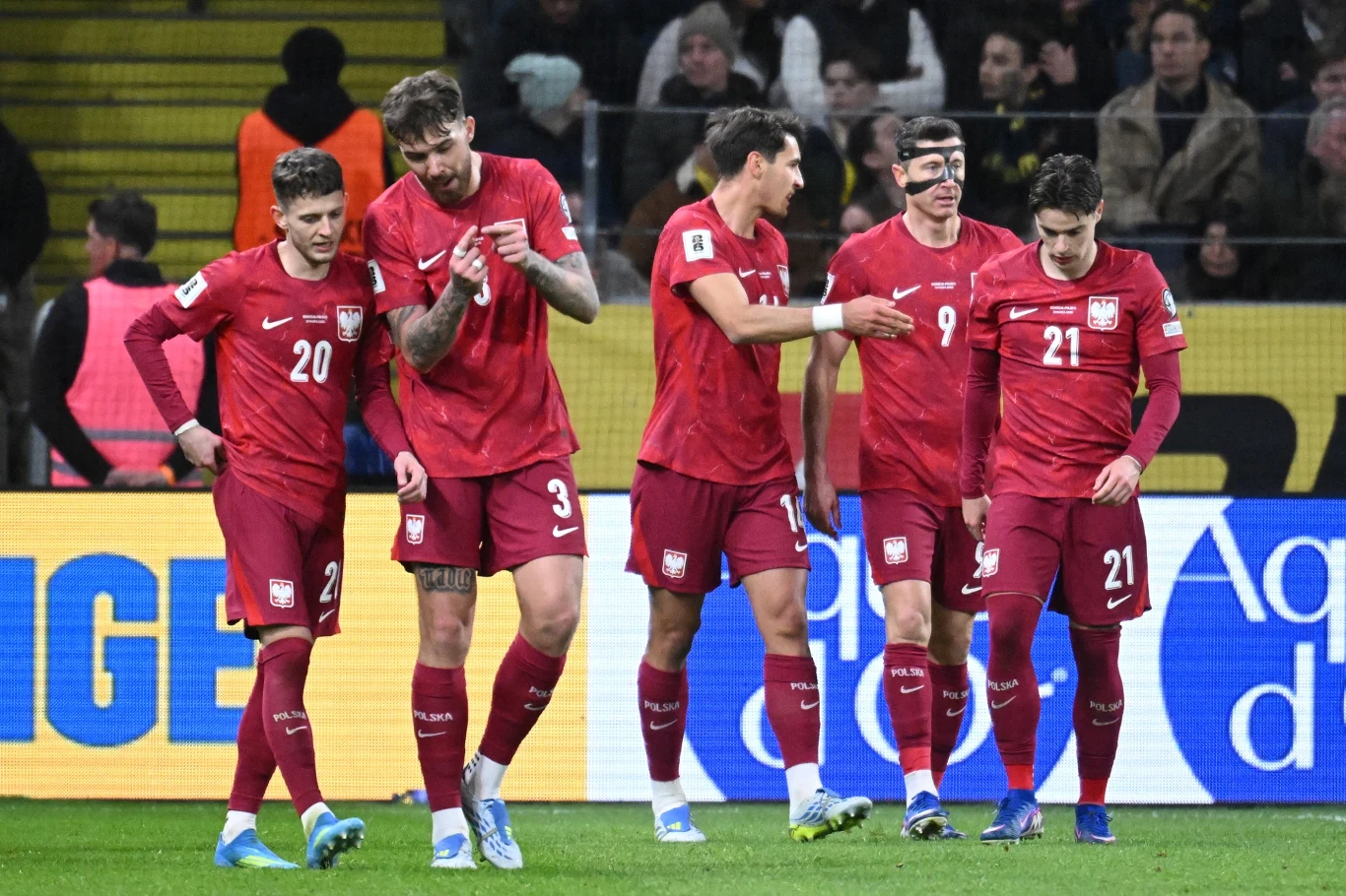 pięciu piłkarzy reprezentacji Polski w czerwonych strojach na murawie, jeden z nich nosi ochronną maskę na twarzy, wokół nich stadionowe otoczenie i kibice na trybunach