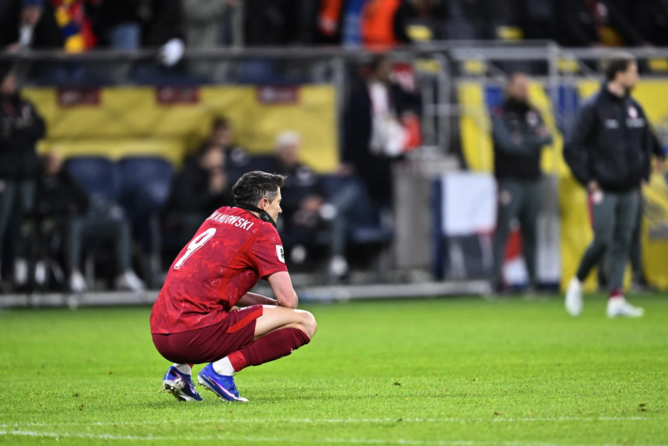 piłkarz w czerwonym stroju z numerem 9 i napisem „Lewandowski” kuca na murawie boiska, wokół niego rozmyte sylwetki osób przy linii bocznej oraz trybuny w tle