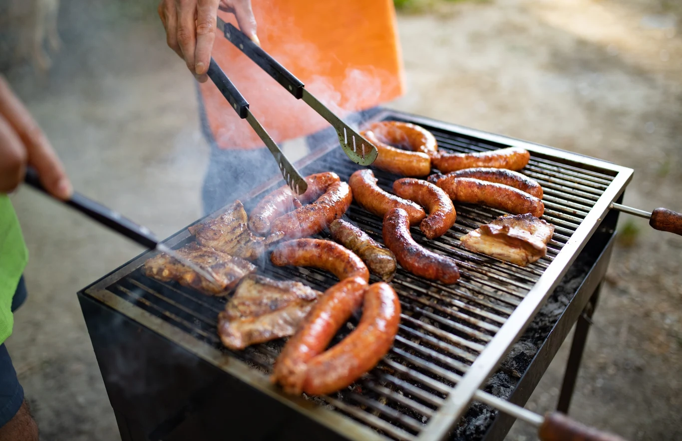 Kiełbaski i kawałki mięsa grillowane na ruszcie, unoszący się dym, ręka w pomarańczowej koszulce obracająca jedzenie za pomocą szczypiec.