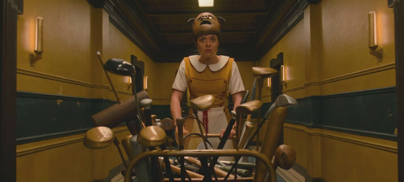 osoba ubrana w żółty strój z białym kołnierzykiem oraz charakterystyczną czapką z niedźwiedzim motywem pcha wózek wypełniony różnorodnymi kijami i rakietami sportowymi w wąskim, żółto-niebieskim korytarzu