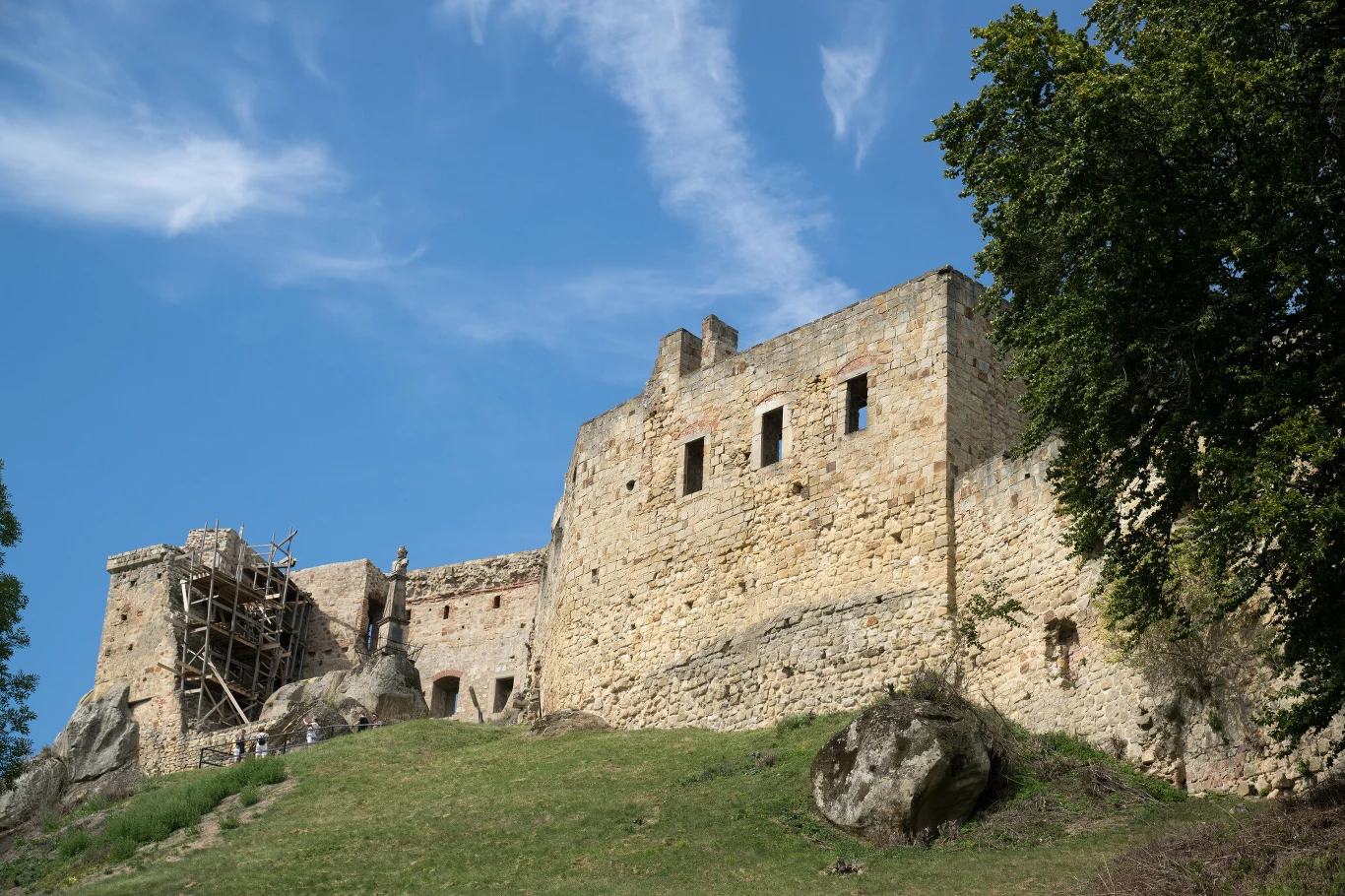 Kamienny zamek na wzgórzu z widocznymi fragmentami murów, otoczony zielenią i dużymi głazami, z częściowo odrestaurowaną konstrukcją po lewej stronie.
