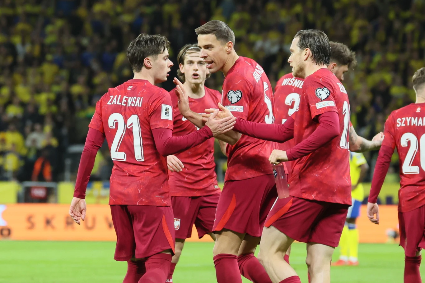 piłkarze polskiej reprezentacji w czerwonych strojach celebrują razem na boisku podczas meczu, wyrażając emocje gestami i mimiką; w tle żółte trybuny z kibicami.