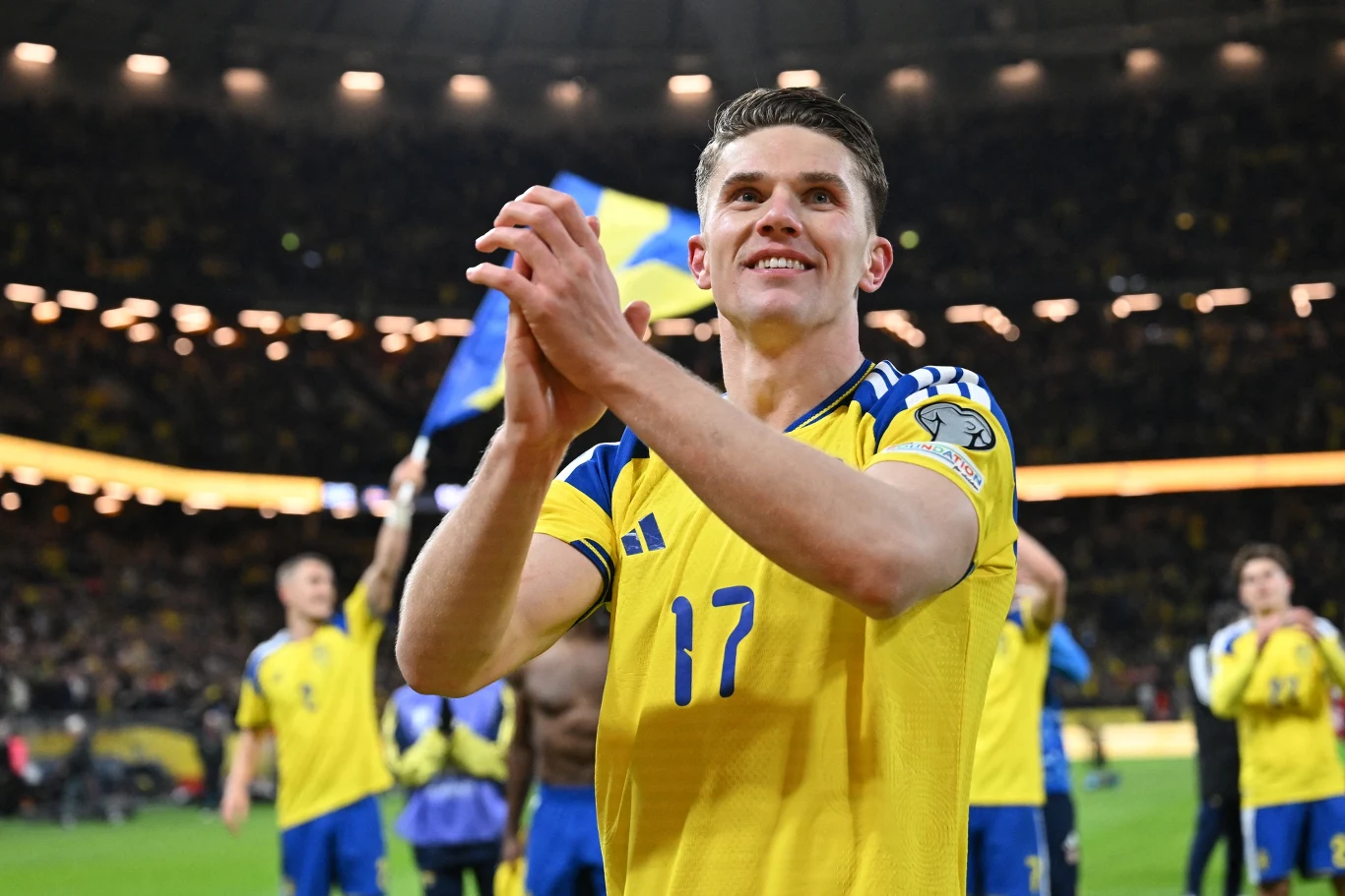 piłkarz w żółtej koszulce z numerem 17 stoi na stadionie, klaszcze w dłonie, w tle kilku innych zawodników świętujących z flagą narodową oraz trybuny wypełnione kibicami