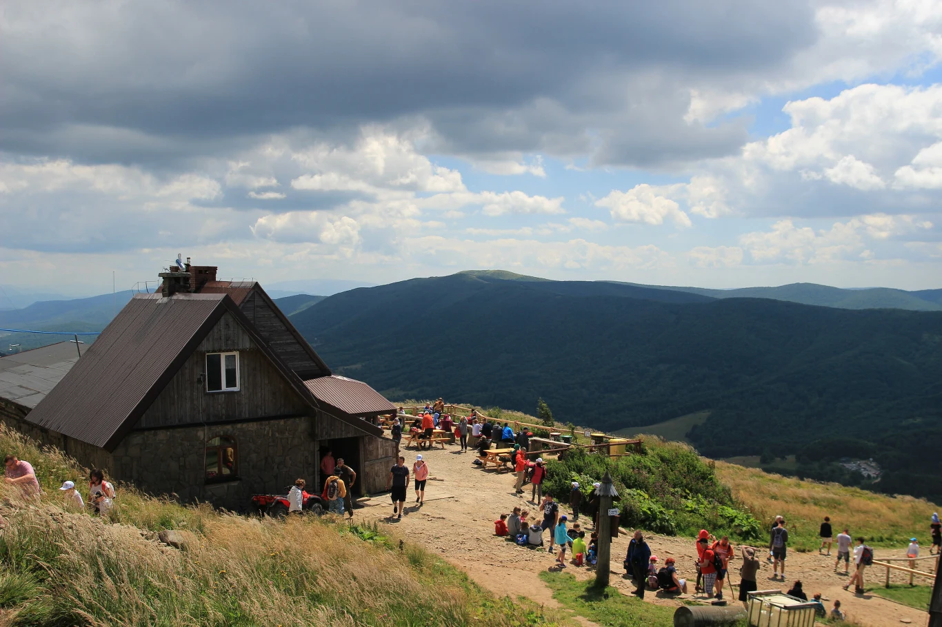 drewniane schronisko górskie otoczone grupami turystów odpoczywających na ławeczkach i trawie, rozległe widoki na zielone wzgórza i ciemne chmury unoszące się nad górami