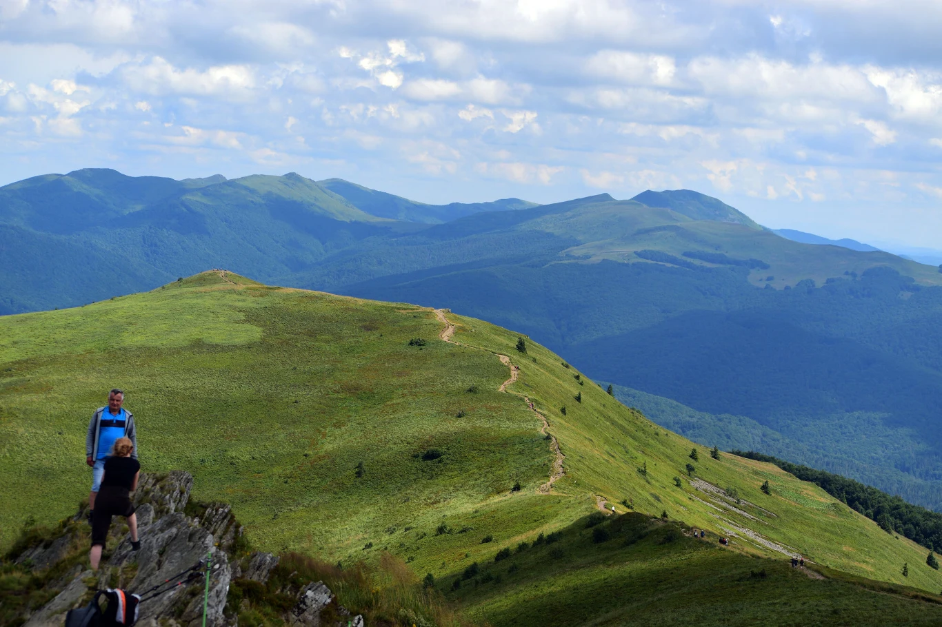 Zielone wzgórza i szlak prowadzący przez rozległe połoniny, po których spacerują turyści, a w tle rozciąga się pasmo górskie pod błękitnym, lekko zachmurzonym niebem.