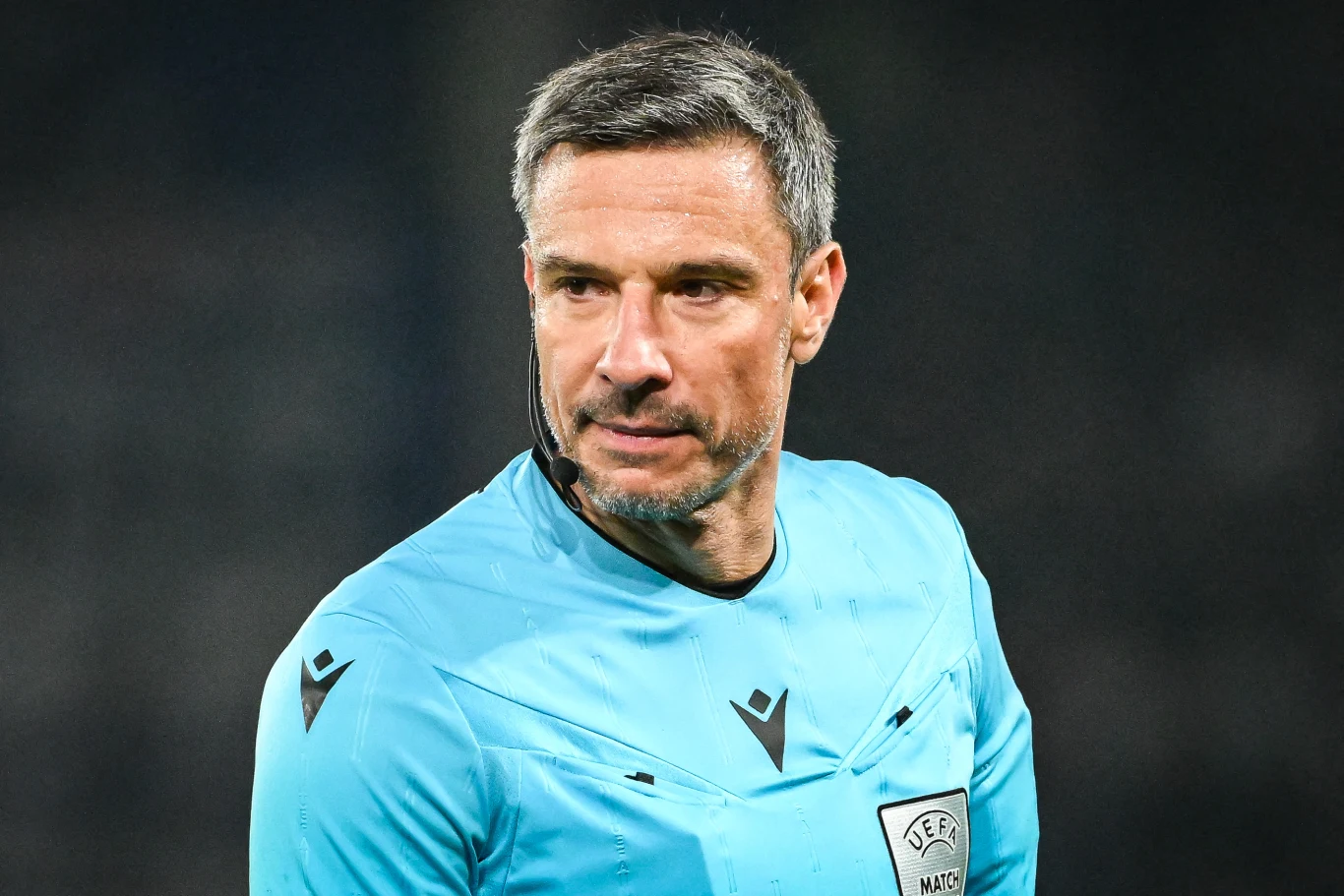 sędzia piłkarski w niebieskim stroju z logotypem UEFA na piersi i mikrofonem przy twarzy, skupiony wzrok skierowany w bok