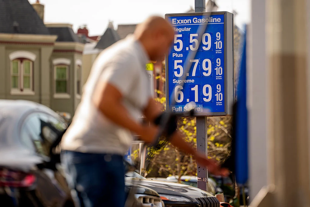 tablica cenowa stacji benzynowej Exxon Gasoline z cenami paliwa na poziomie powyżej 5 dolarów za galon, w tle rozmazana postać tankującej osoby oraz fragmenty zaparkowanych samochodów