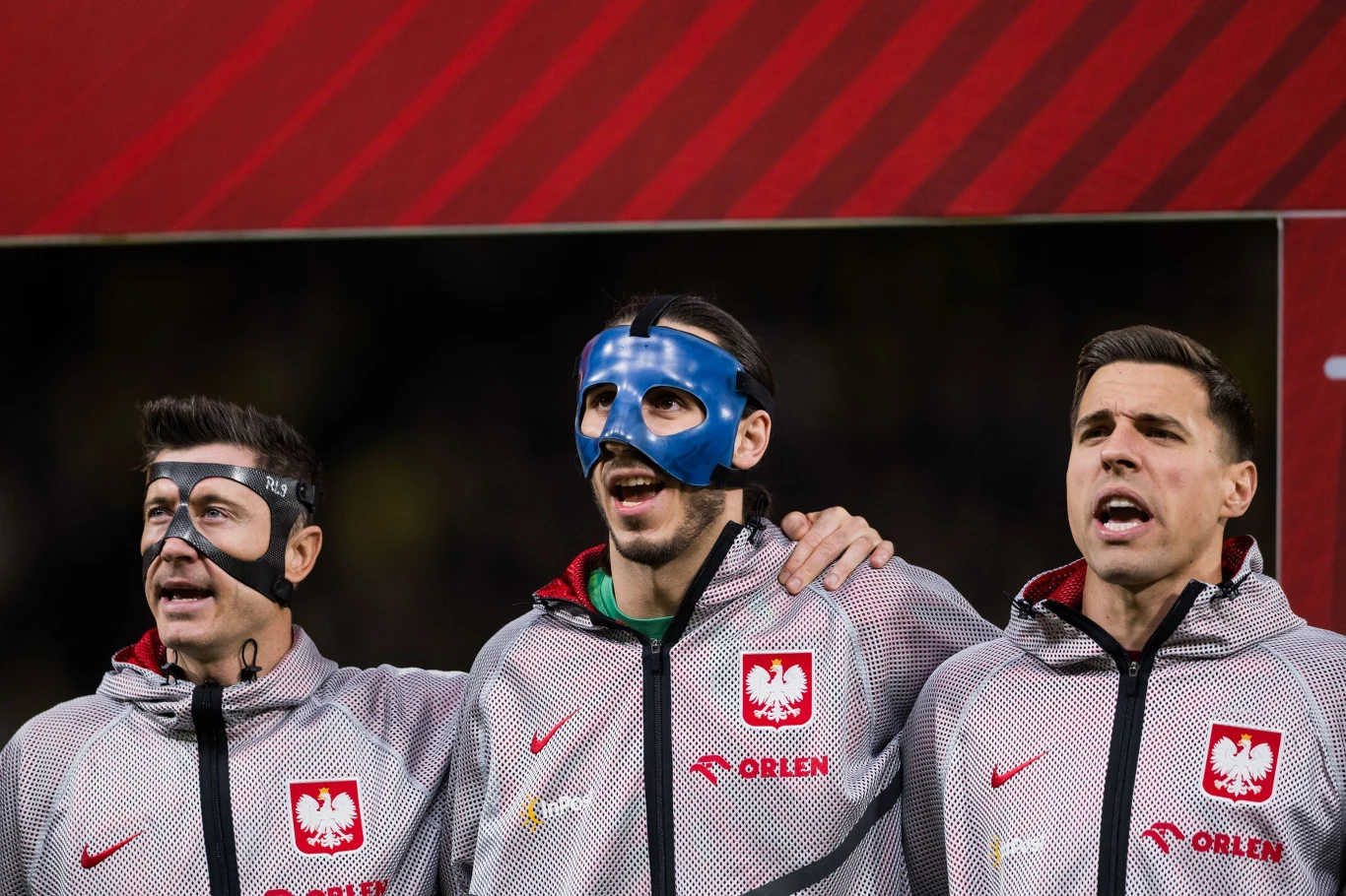 trzech polskich piłkarzy w reprezentacyjnych dresach z herbem Polski śpiewających hymn, dwóch z nich nosi ochronne maski na twarzy