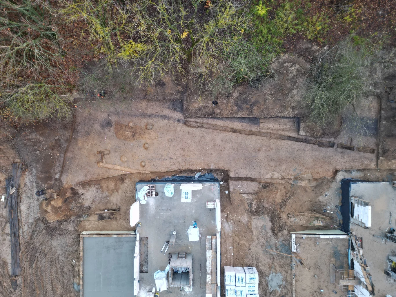 teren wykopalisk archeologicznych z odsłoniętą drogą przebiegającą przez środek obrazu, otoczoną ziemią i fragmentami zabudowy, w tle drzewa i bujna roślinność