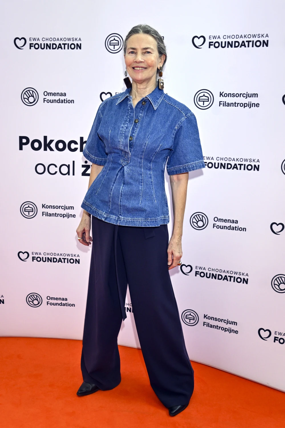 kobieta w średnim wieku uśmiecha się, ubrana w niebieską koszulową bluzkę z krótkimi rękawami oraz szerokie, ciemne spodnie, stojąc na pomarańczowym dywanie na tle ścianki z logotypami fundacji i sponsorów