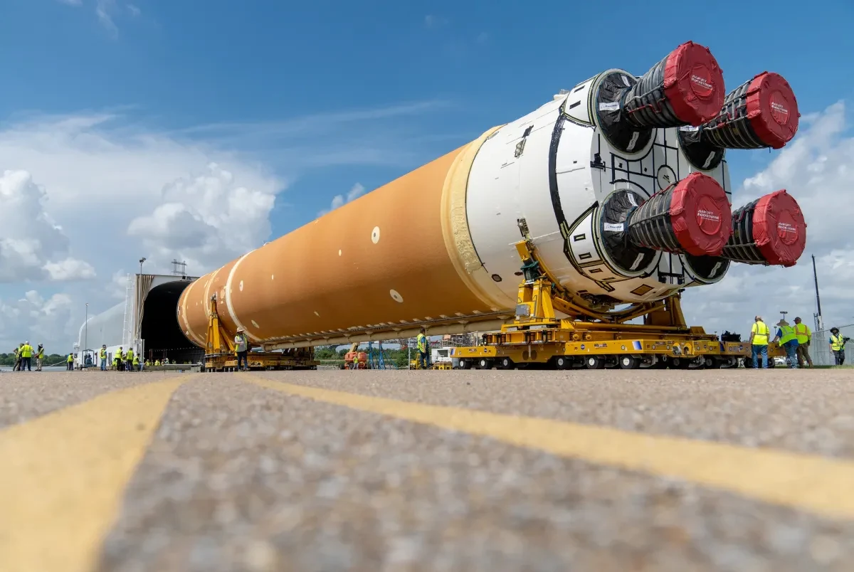 Ogromny, cylindryczny segment rakiety transportowany jest na specjalnej platformie przez zespół techników w żółtych kamizelkach na tle nieba z białymi chmurami.
