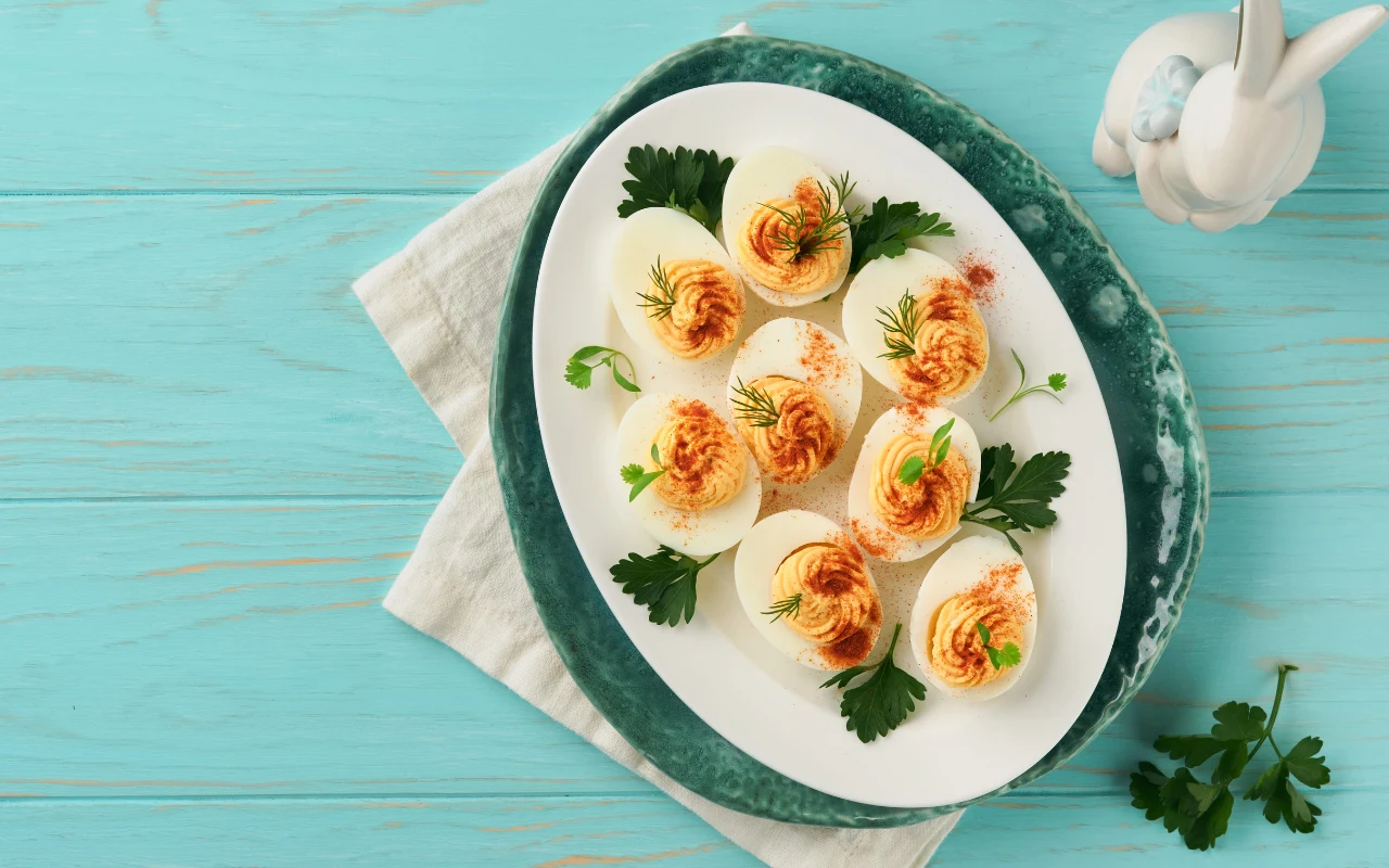 Faszerowane jajka to zdrowa propozycja z wielkanocnego stołu Połówki jajek faszerowanych kremowym nadzieniem, posypane papryką i udekorowane gałązkami koperku oraz natką pietruszki, ułożone na białym półmisku na turkusowym stole obok figurki białego królika.