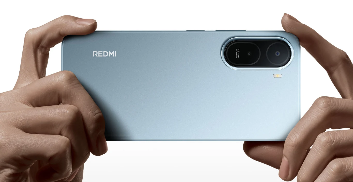 Srebrny smartfon marki Redmi trzymany poziomo oburącz, z wyeksponowanym podłużnym obiektywem aparatu oraz logo REDMI po lewej stronie obudowy.