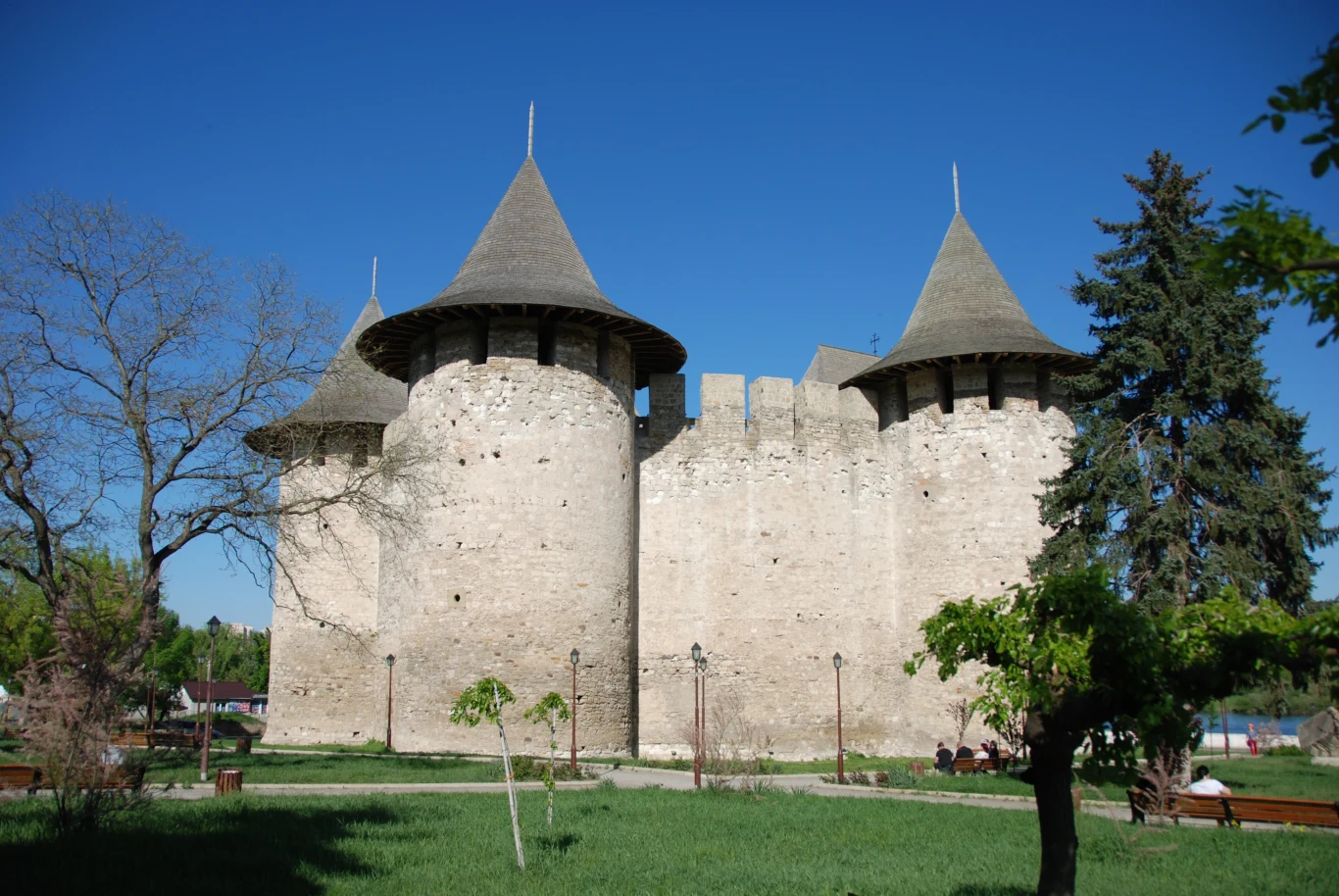 masywne, kamienne mury zamku z trzema wysokimi, okrągłymi wieżami o stożkowych dachach, otoczone zielenią oraz rzadkimi drzewami