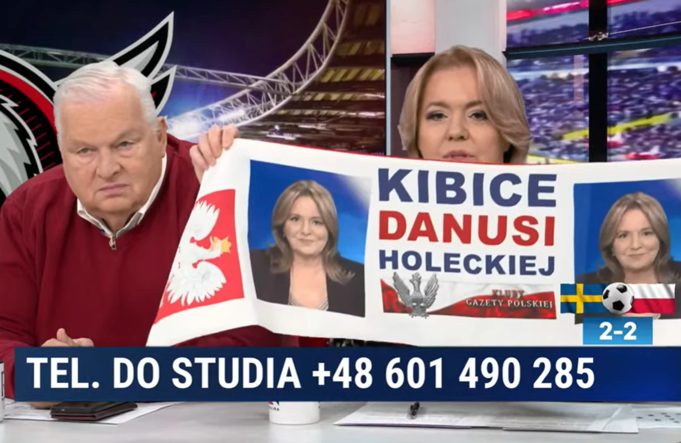 Dwoje prowadzących w studiu telewizyjnym trzyma transparent z napisem „Kibice Danusi Holeckiej” i jej zdjęciami, na dole ekranu widoczny numer telefonu do studia oraz grafika z wynikiem meczu Polska-Szwecja 2-2.