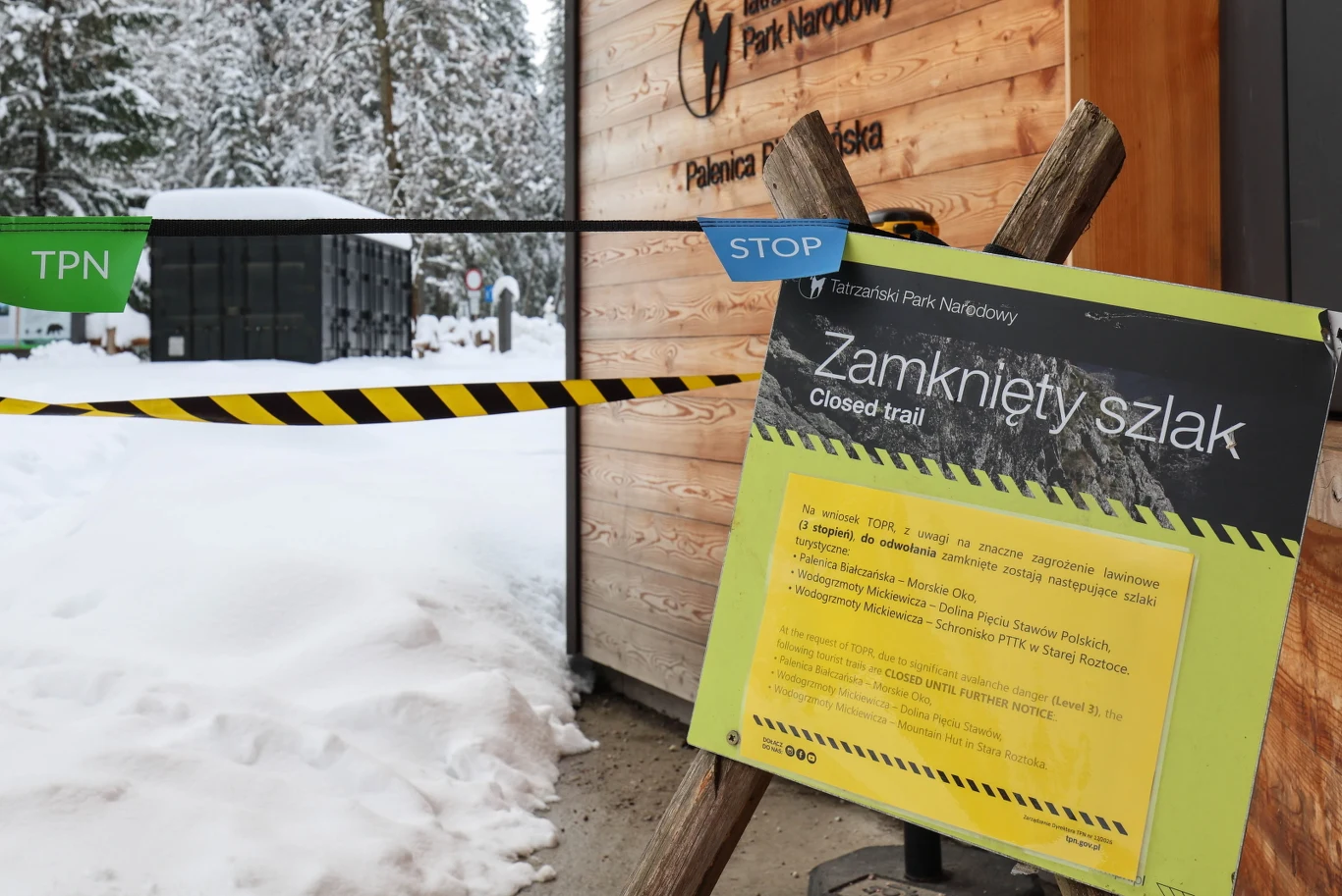 Zaklejona taśmą i oznakowana tablicą z napisem „Zamknięty szlak” wejście do ośnieżonego Tatrzańskiego Parku Narodowego, obok drewniany budynek i zasypany śniegiem teren leśny w tle.