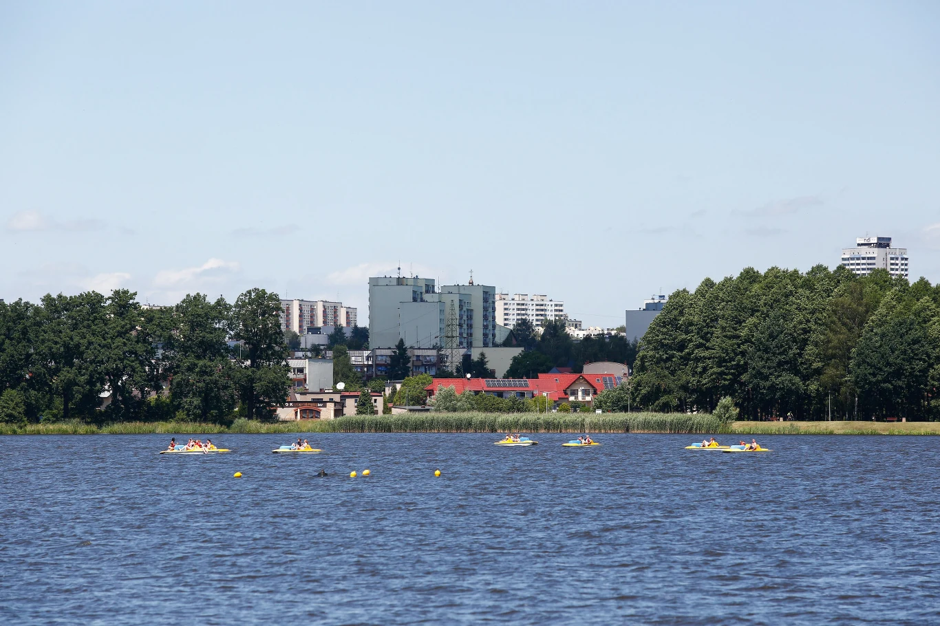 Kilka rowerów wodnych pływa po jeziorze, w tle widoczne są budynki mieszkalne oraz otoczenie zielonych drzew i niskich zabudowań przy brzegu.