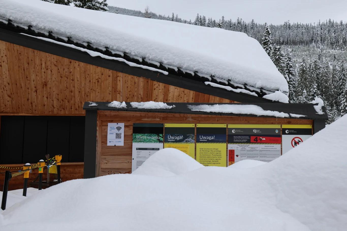 drewniany budynek pokryty grubą warstwą śniegu, tablice informacyjne umieszczone na ścianie oraz wysokie zaspy śniegu przed wejściem; w tle widać zaśnieżone drzewa