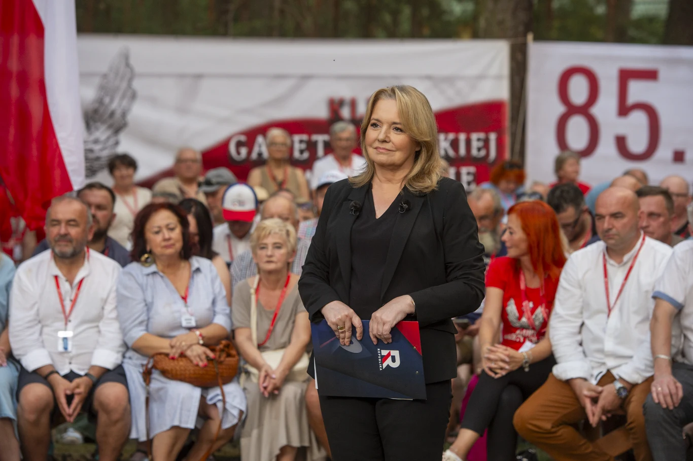 kobieta w czarnym garniturze stoi na tle siedzących uczestników wydarzenia plenerowego; w tle widoczne biało-czerwone elementy dekoracyjne oraz banner z napisem i liczbą 85