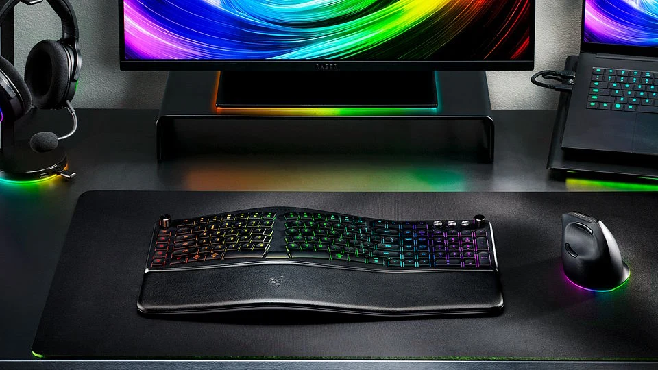 Ergonomiczna klawiatura z podświetleniem RGB umieszczona na dużej podkładce, obok bezprzewodowa myszka również z podświetleniem, monitor z kolorowym ekranem oraz zestaw słuchawkowy na stojaku w tle.