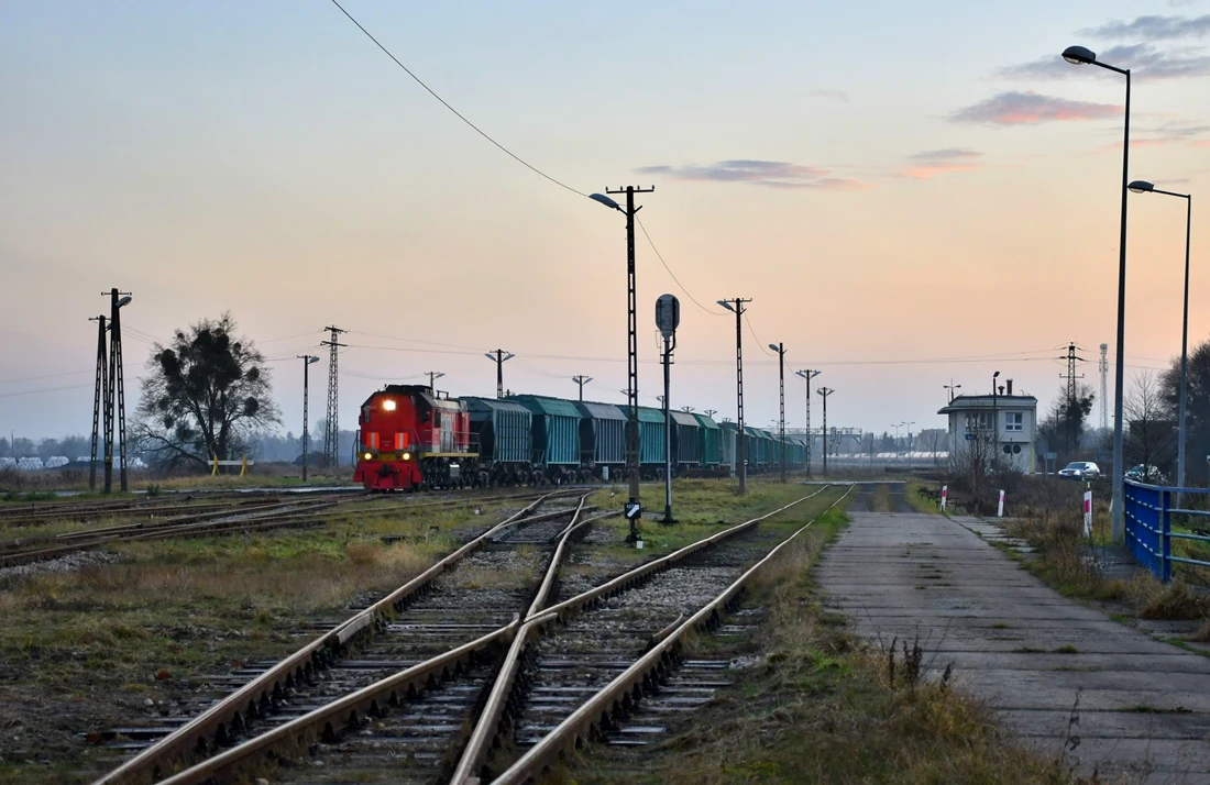 Czerwony lokomotywa z ciągiem zielonych wagonów towarowych jadąca po torach kolejowych na rozjeździe, obok starej nastawni i otoczenia industrialnego o zmierzchu.
