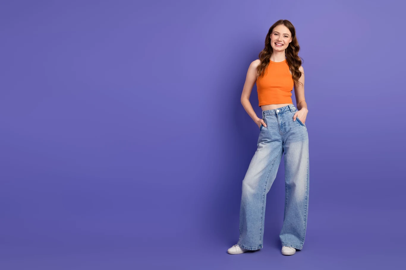młoda kobieta z długimi, falowanymi włosami ubrana w pomarańczowy top i szerokie dżinsy, stojąca na fioletowym tle z rękami w kieszeniach i szerokim uśmiechem