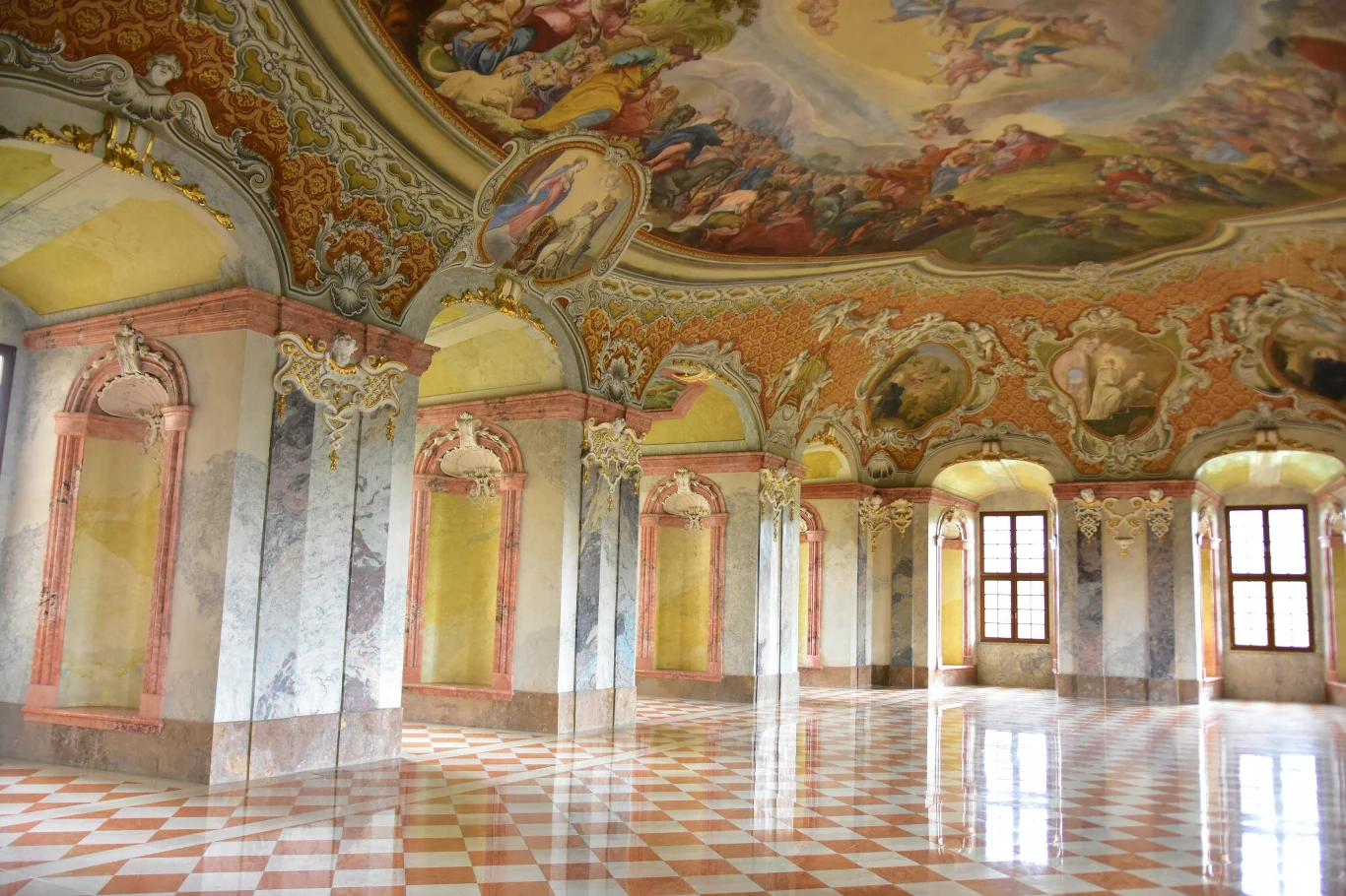 bogato zdobiona sala barokowa z marmurowymi kolumnami, malowidłami ściennymi oraz sufitowymi przedstawiającymi sceny mitologiczne, błyszcząca posadzka w biało-różową szachownicę, duże, łukowate okna wpuszczające naturalne światło