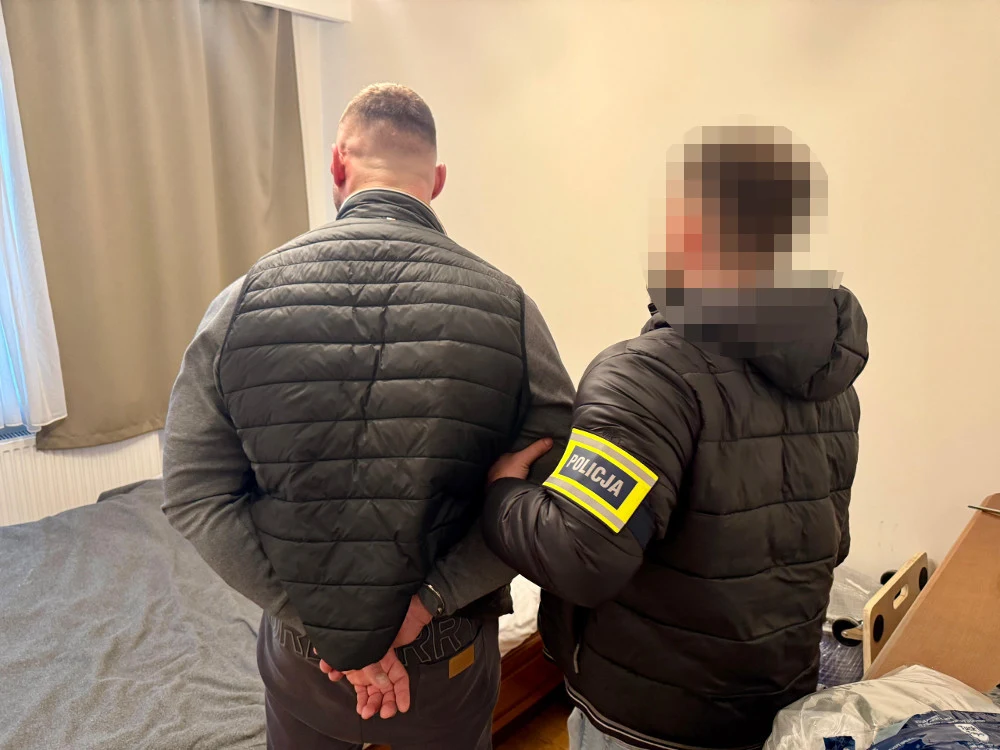 policjant w czarnej kurtce z widoczną naszywką 'policja' na rękawie zatrzymuje mężczyznę z rękami skute kajdankami za plecami, obaj stoją w pokoju z łóżkiem i zasłoniętym oknem