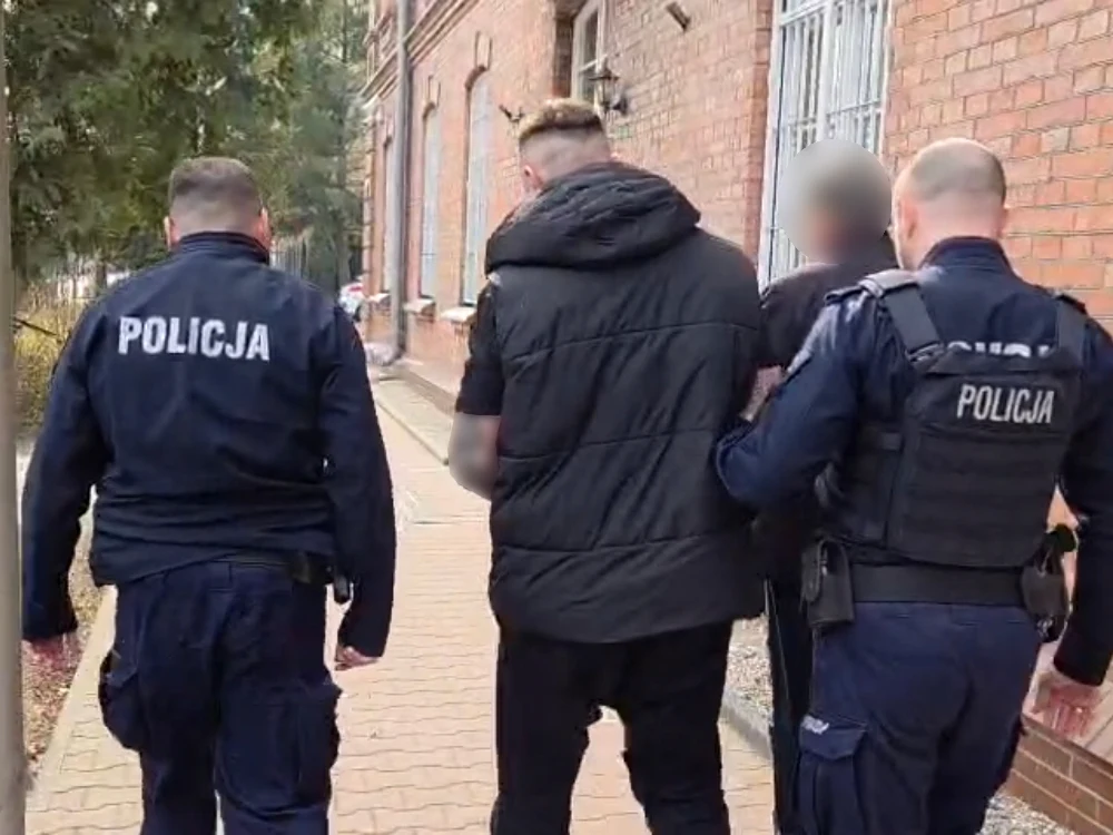 dwóch policjantów eskortuje mężczyznę o zamazanej twarzy wzdłuż chodnika przy ceglanej kamienicy
