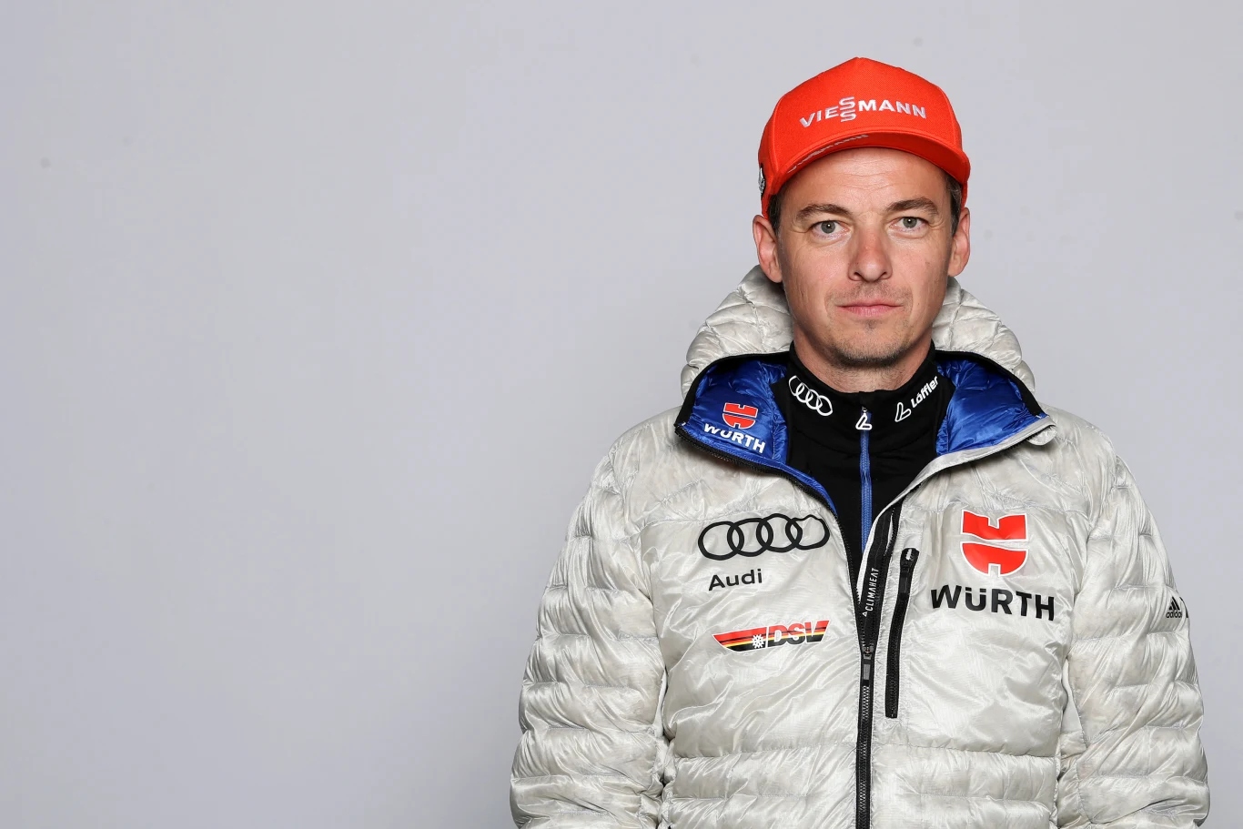mężczyzna w sportowej kurtce z logotypami Würth oraz Audi, ubrany w czerwoną czapkę z napisem Viessmann, stoi na jasnym tle, patrząc prosto w obiektyw